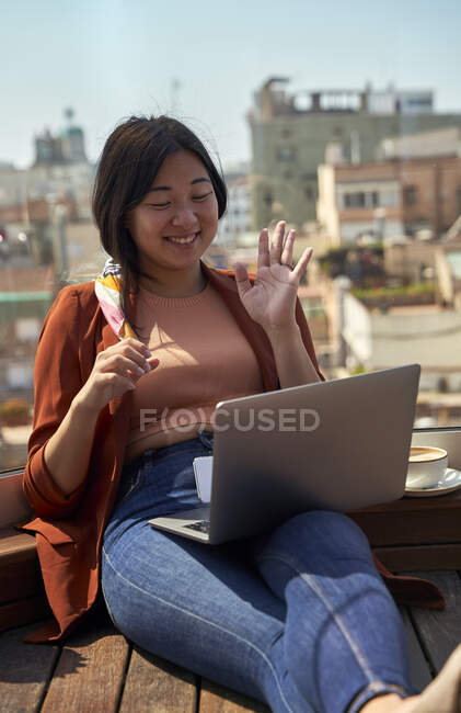 Усміхнена жінка махає рукою до відеодзвінка на ноутбуці, сидячи на терасі даху. — стокове фото