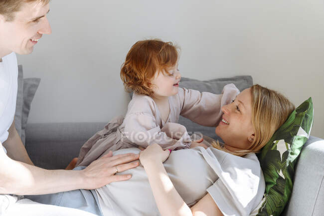 Fofa filha admirar grávida mãe por pai no sofá em casa — Fotografia de Stock