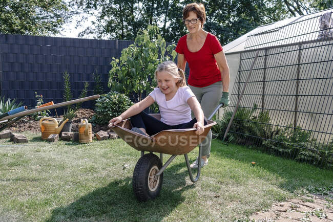 Glückliches Mädchen sitzt in Schubkarre beim Spielen mit Großmutter im Hinterhof — Stockfoto