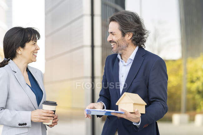 Smiling male real estate agent with house model discussing with female customer holding disposable cup in park — Stock Photo