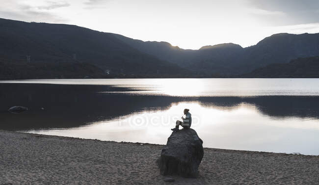 Uomo seduto sulla roccia vicino al lago durante il tramonto — Foto stock