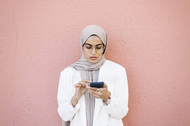 Jeune femme utilisant un téléphone intelligent tout en se tenant devant le mur rose — Photo de stock