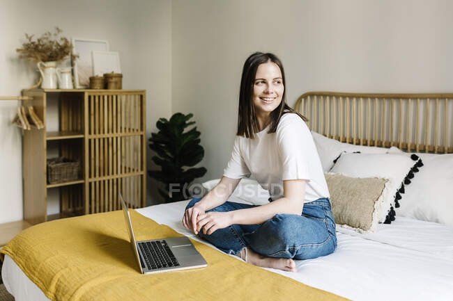 Mujer sonriente sentada con portátil en la cama en casa - foto de stock