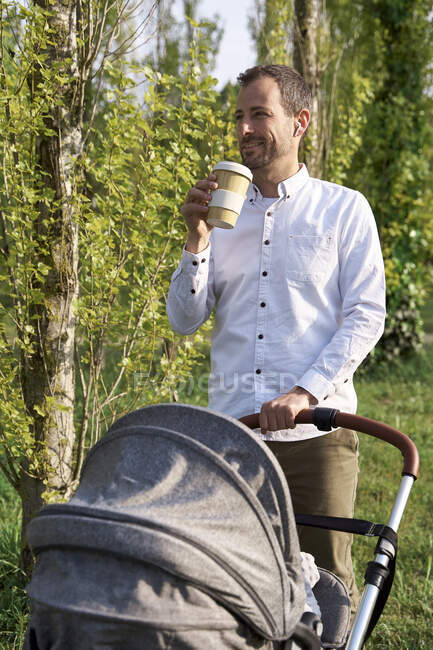 Homme adulte moyen buvant du café debout avec une poussette bébé le jour ensoleillé — Photo de stock
