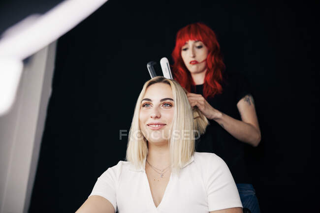 Bella modello femminile sorridente mentre parrucchiere facendo raddrizzamento davanti alla luce anello — Foto stock