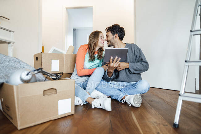 Feliz pareja joven con la tableta sentada junto a cajas de cartón en el nuevo hogar - foto de stock