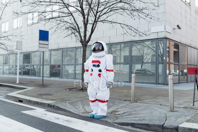 Joven astronauta en traje espacial de pie cerca del cruce de cebra fuera del edificio en la ciudad - foto de stock