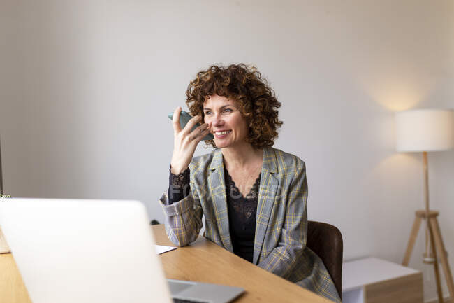 Усмішка жіночого фрилансеру посилає голосове повідомлення через смартфон, працюючи вдома. — стокове фото