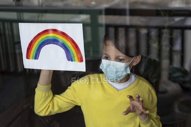 Мальчик смотрит, держа радугу на бумаге на стекле в защитной маске для лица во время COVID-19 — стоковое фото