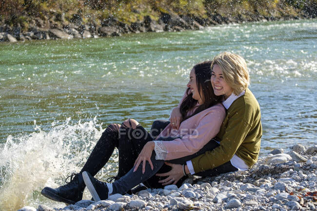 Веселая лесбийская пара, сидящая на берегу озера, пока вода брызгает на камни в солнечный день — стоковое фото