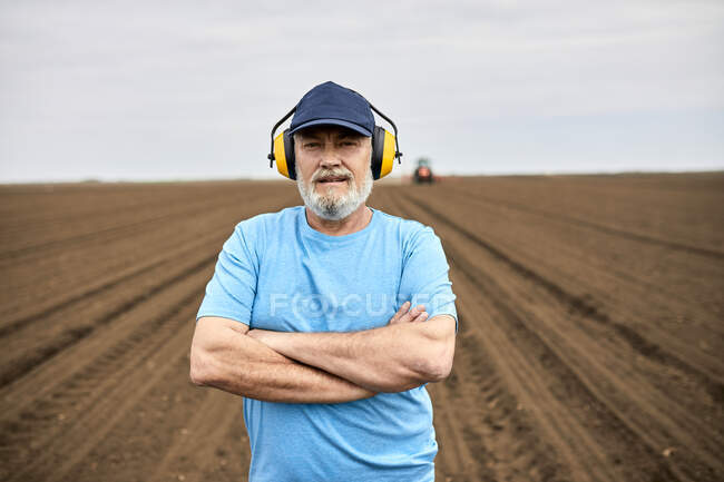 Мужчина-фермер носит защитные ушки, стоя с оружием, скрещенным на сельскохозяйственном поле — стоковое фото