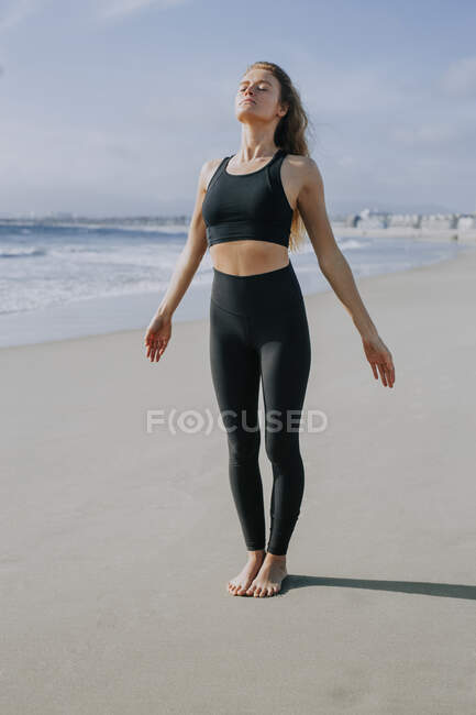 Jovem mulher em roupas esportivas de pé com os olhos fechados na praia no dia ensolarado — Fotografia de Stock