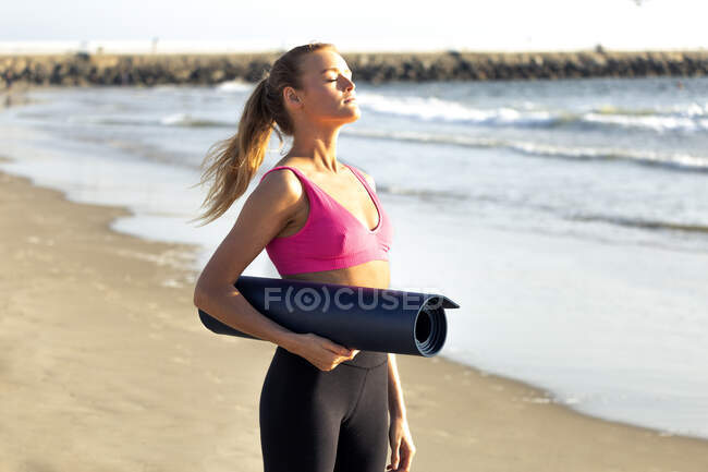 Mulher bonita segurando tapete de exercício enquanto de pé com os olhos fechados na praia — Fotografia de Stock