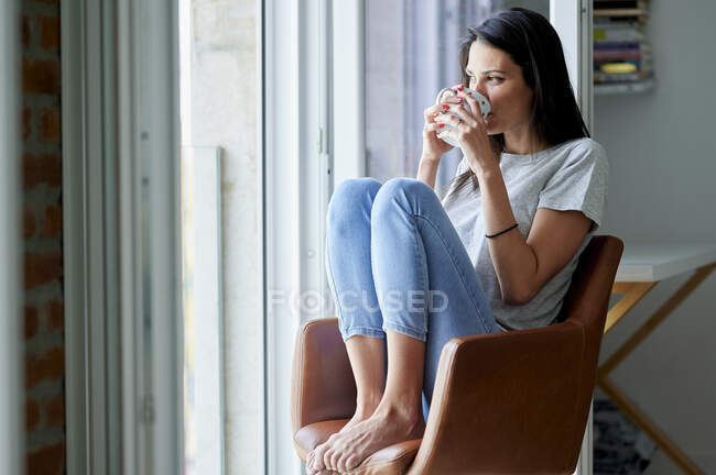 Mujer reflexiva mirando hacia otro lado mientras bebe café en casa - foto de stock