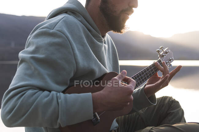 Uomo con camicia incappucciata giocare ukulele al lago — Foto stock