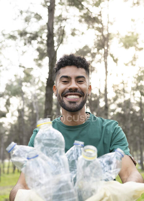 Joven voluntario sonriente recogiendo botellas de plástico en el bosque - foto de stock