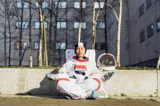 Female astronaut meditating while sitting by retaining wall during sunny day — Stock Photo