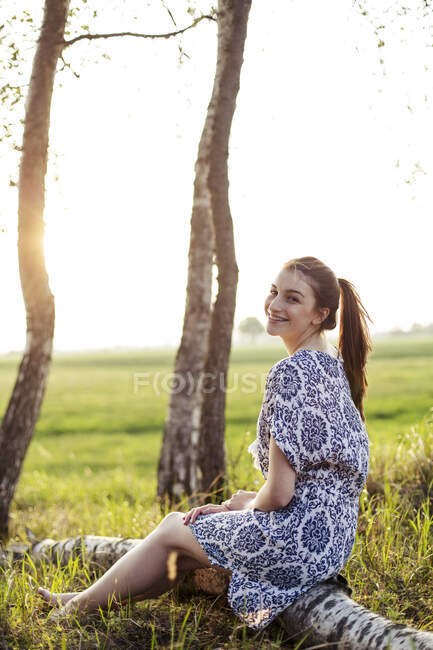 Mujer joven sonriente sentada en el tronco de un árbol y disfrutando del atardecer - foto de stock