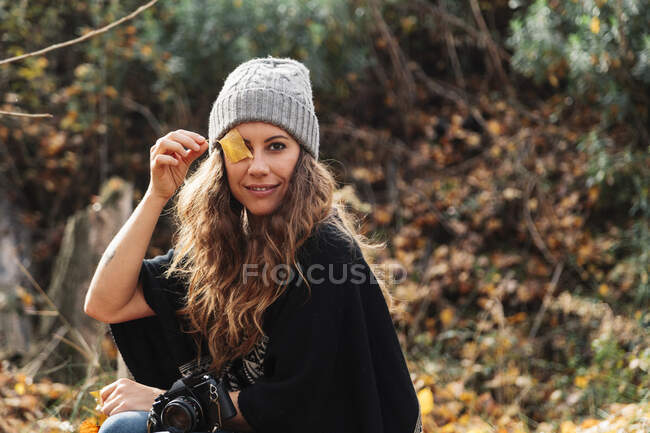 Smiling woman covering eye with autumn leaf while sitting in forest during vacations — Stock Photo