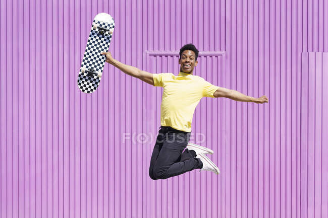 Hombre alegre con los brazos extendidos sosteniendo monopatín entero saltando en hierro ondulado púrpura - foto de stock