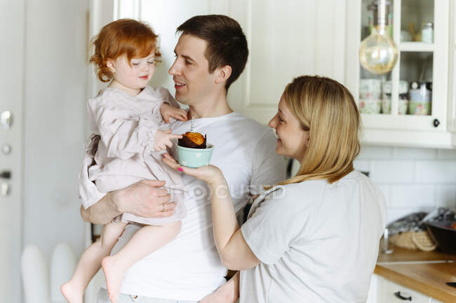 Madre sorridente che dà cupcake alla figlia portata dal padre in cucina a casa — Foto stock