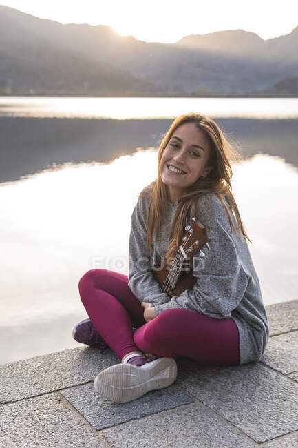 Donna sorridente con ukulele seduta a gambe incrociate sul lungomare vicino al lago — Foto stock