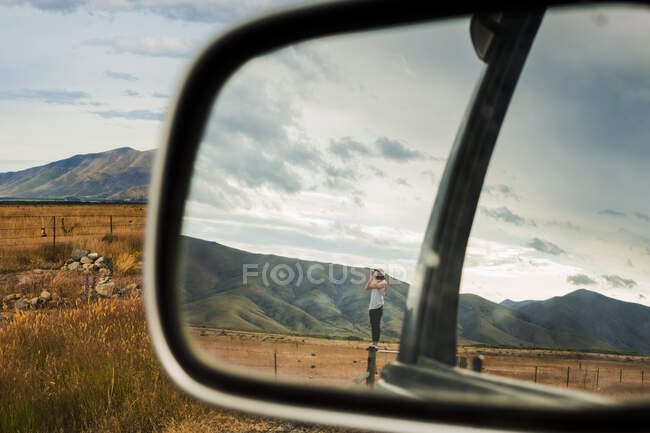Spiegelbild eines jungen Mannes, der auf einem Zaun steht und durch ein Fernglas auf die umliegenden Berge blickt — Stockfoto