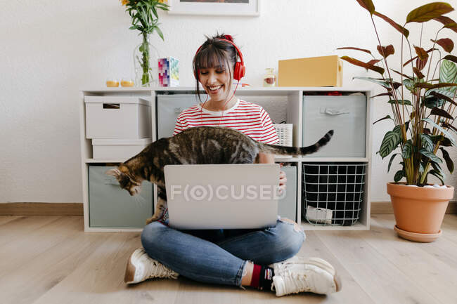 Giovane donna che indossa cuffie seduta con computer portatile e gatto a casa — Foto stock