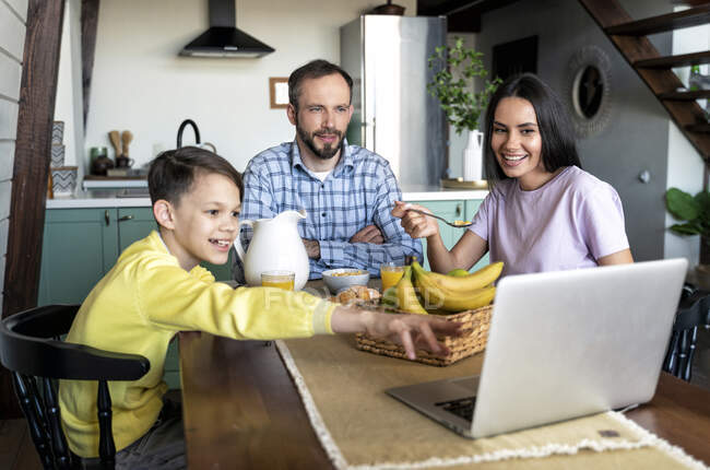 Familia mirando a la computadora portátil mientras desayuna en la mesa de comedor en casa - foto de stock