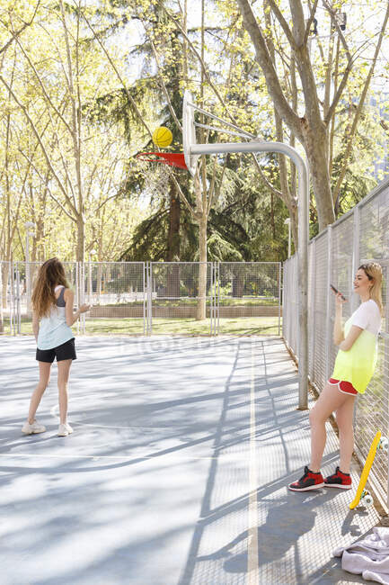 Young woman talking on mobile phone with friend playing basketball in sports court — Stock Photo