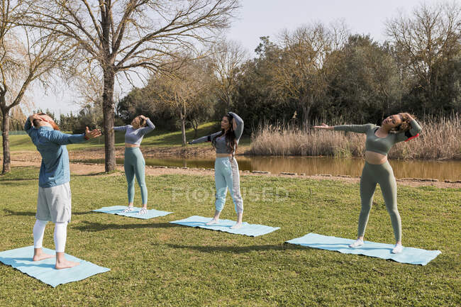Мужской инструктор по йоге, направляющий молодых женщин во время упражнений на растяжку на коврике в солнечный день — Йога, мультикультурный - Stock Photo