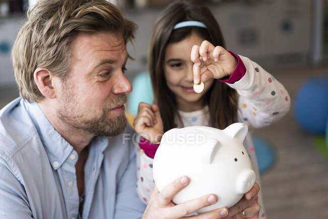 Tochter legt Münze in Sparschwein, das Vater zu Hause hält — Stockfoto
