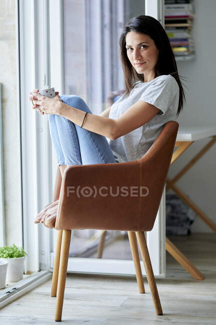 Bella donna in possesso di tazza di caffè mentre seduto sulla sedia a casa — Foto stock