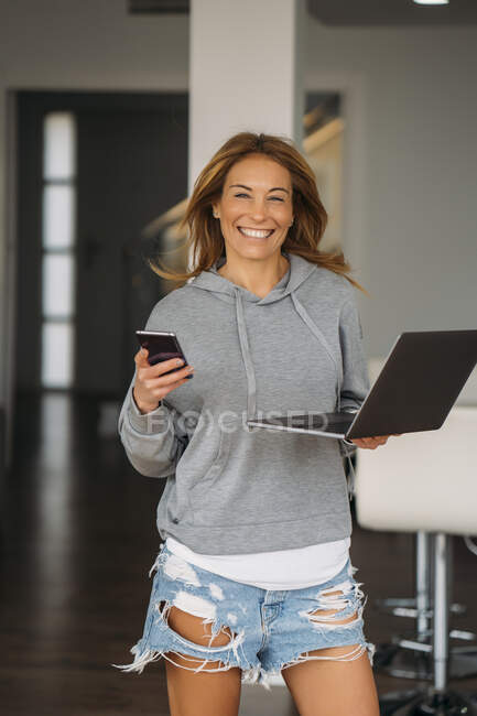 Веселая женщина-профессионал держит ноутбук и смартфон во время прогулки по дому — стоковое фото