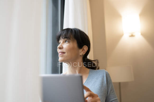 Donna matura guardando attraverso la finestra durante l'utilizzo di tablet digitale in camera d'albergo — Foto stock