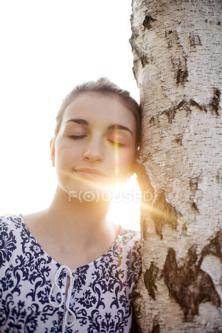 Mujer joven con los ojos cerrados apoyados en un tronco de árbol disfrutando de la naturaleza - foto de stock