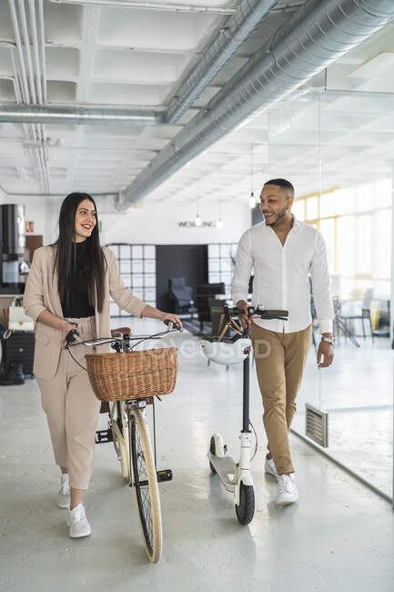 Smiling businessman looking at female colleague while walking with bicycles at coworking office — Stock Photo