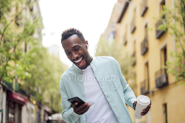 Сміється з молодого чоловіка з чашкою багаторазового використання, користуючись смартфоном на вулиці. — стокове фото