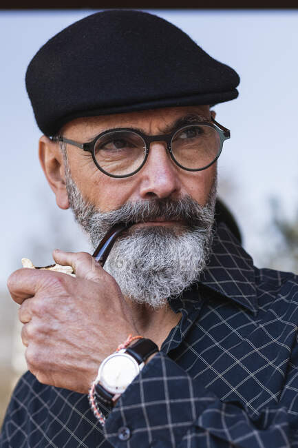 Бородатый мужчина в плоской шляпе смотрит в сторону, держа трубку для курения — стоковое фото