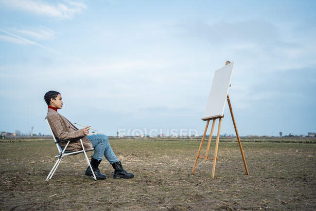 Thoughtful woman looking at easel while sitting on chair in countryside — Stock Photo