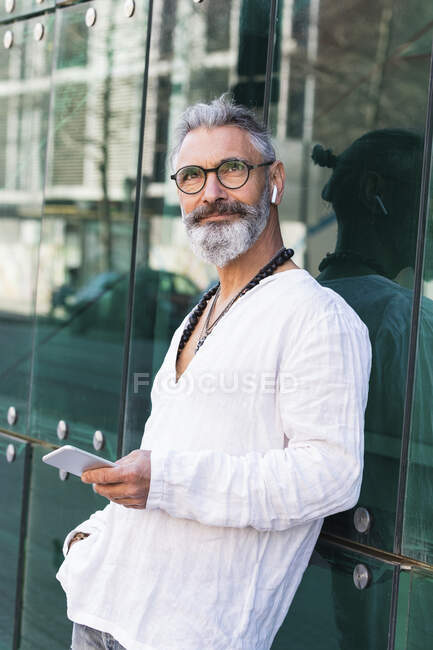 Homme barbu avec téléphone portable rêvant jour tout en s'appuyant sur le mur de verre — Photo de stock