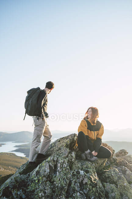 Joven excursionista sentada por el hombre de pie en la cima de la montaña - foto de stock
