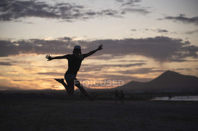 Mujer excitada con los brazos extendidos saltando en la playa al atardecer - foto de stock