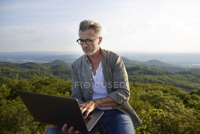 Человек в очках с ноутбуком, сидя на горе — стоковое фото
