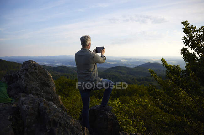 Человек фотографирует пейзаж через цифровую табличку с горы во время заката — стоковое фото