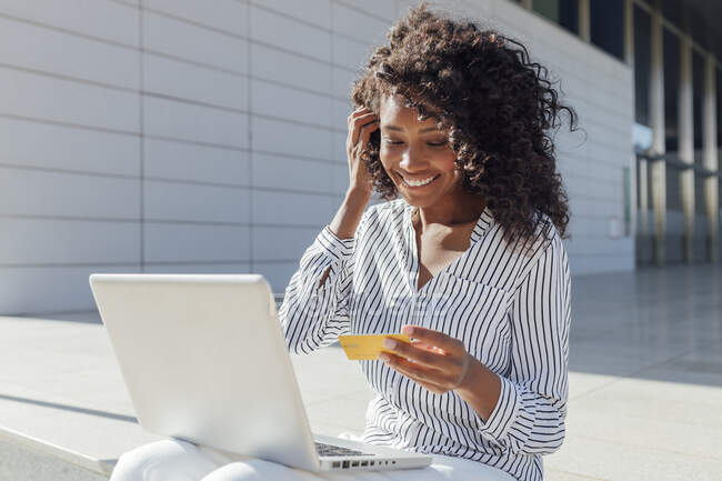 Smiling woman with credit card doing online shopping while sitting outside office building — Stock Photo