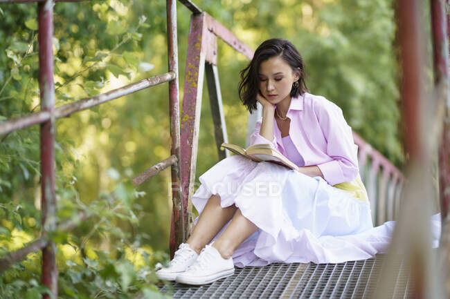 Junge Frau sitzt mit der Hand am Kinn und liest Buch auf Fußgängerbrücke im Grünen — Stockfoto