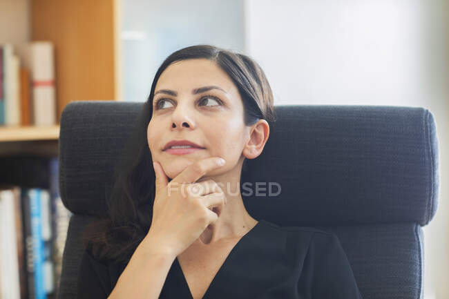 Ragionevole donna d'affari con la mano sul mento distogliendo lo sguardo seduta in ufficio — Foto stock