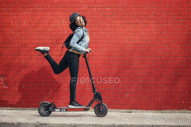 Carefree young woman riding electric push scooter by red brick wall — Stock Photo