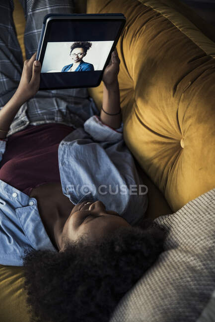 Donna sorridente che parla con un amico attraverso il tablet digitale mentre è sdraiato sul divano — Foto stock